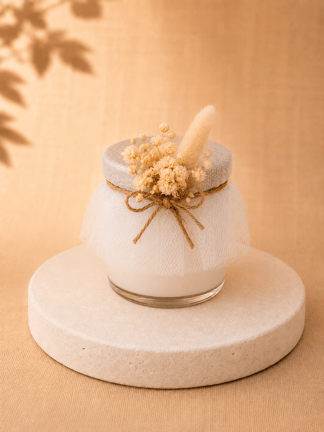 White wedding candle favour wrapped in tulle and decorated with beige dried flowers, displayed on a beige pedestal.